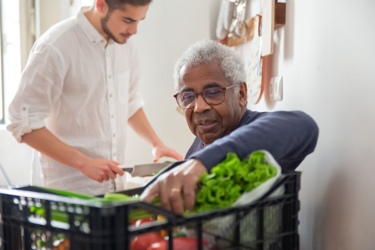 A volunteer supporting an elderly man carrying groceries